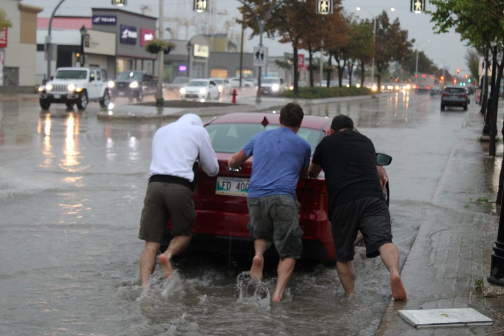 GREG VANDERMEULEN THE CARILLON
These trio of good Samaritans had no problem stepping into the driving rain, removing their footware and helping out a friend in need. This car was one of many that stalled out in the deep water present on Main Street near Brandt Street on Tuesday afternoon.