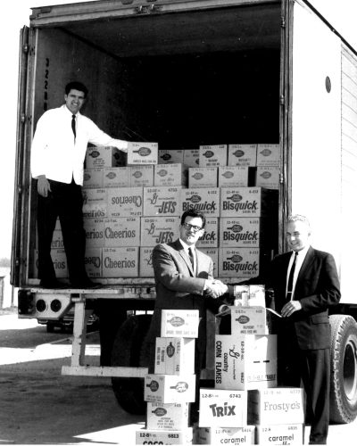 CARILLON ARCHIVES 

Ken Walker of General Mills and Jim Penner of Penner’s Tom-Boy with stacks of cases of General Mills cereal and Betty Crocker cake mixes, part of an 1,800 case shipment, being unloaded under the supervision of Gil Gerbrandt at the Steinbach store.